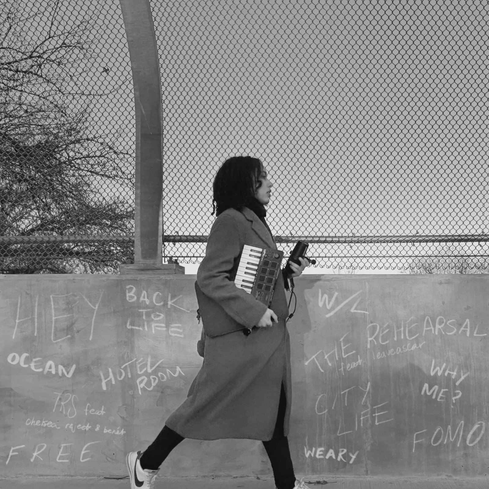 Aja Adam walking with a MIDI keyboard and a camera past a wall chalked with the Quatre track titles. Hero photo from ajaadam.com.